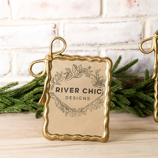 Decorative ornament with 'River Chic Designs' branding on a wooden surface with greenery.