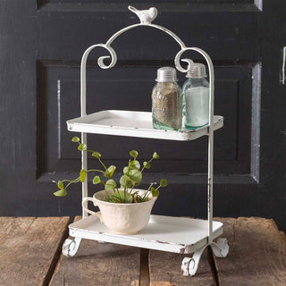 A white two-tiered metal tray with a bird design on the top, mounted on wheels, displaying plants and jars on its tiers.