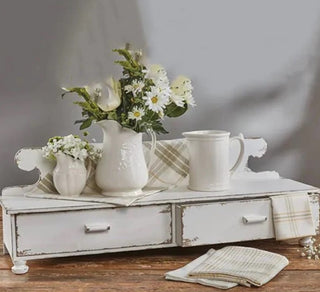 A distressed white wooden counter shelf with two drawers, decorated with a vase and floral arrangement on top, alongside a cup and saucer.