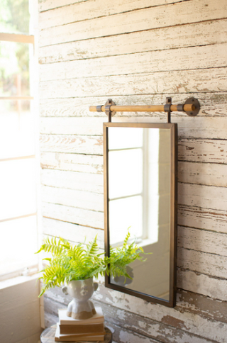 A rectangular wall mirror with a wooden dowel hanger mounted on a wall, with a plant and decorative items placed to its bottom left.