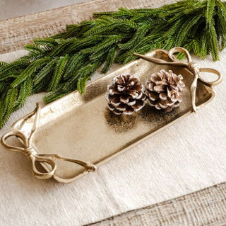 Decorative tray with gold handles, pinecones, and greenery on a textured surface