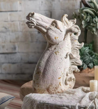 A beige magnesia horsehead sculpture with a textured finish, placed on a surface with a brick wall and greenery in the background.
