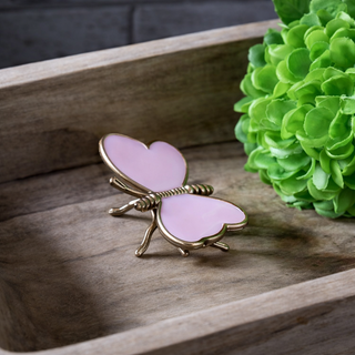 Butterfly-shaped object with pink wings and gold frame on a wooden surface with green foliage in the background