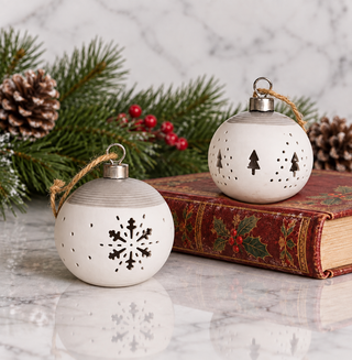 Two white ceramic Christmas ornament balls with laser-cut holiday designs, one featuring a snowflake and the other a tree pattern, displayed on a vintage red book with pine branches and red berries in the background.