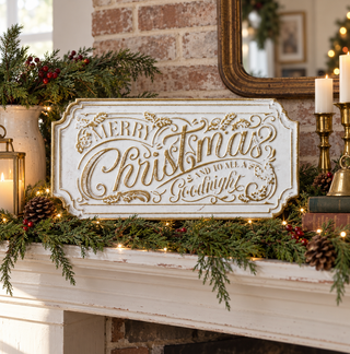 A distressed white metal Christmas sign with gold lettering reading 'Merry Christmas and to All a Good Night' displayed on a decorated fireplace mantel with green garlands, pinecones, candles, and warm string lights.