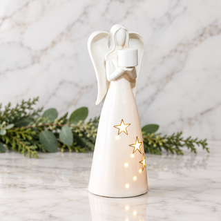 A white ceramic angel figurine with gold star cutouts, holding a small white cup, standing on a marble surface with green foliage and a marble wall backdrop.