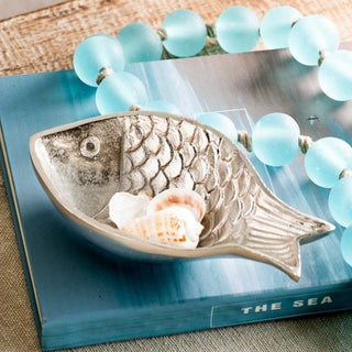 Decorative fish-shaped bowl with a shell on a book titled 'The Sea', surrounded by blue beads.